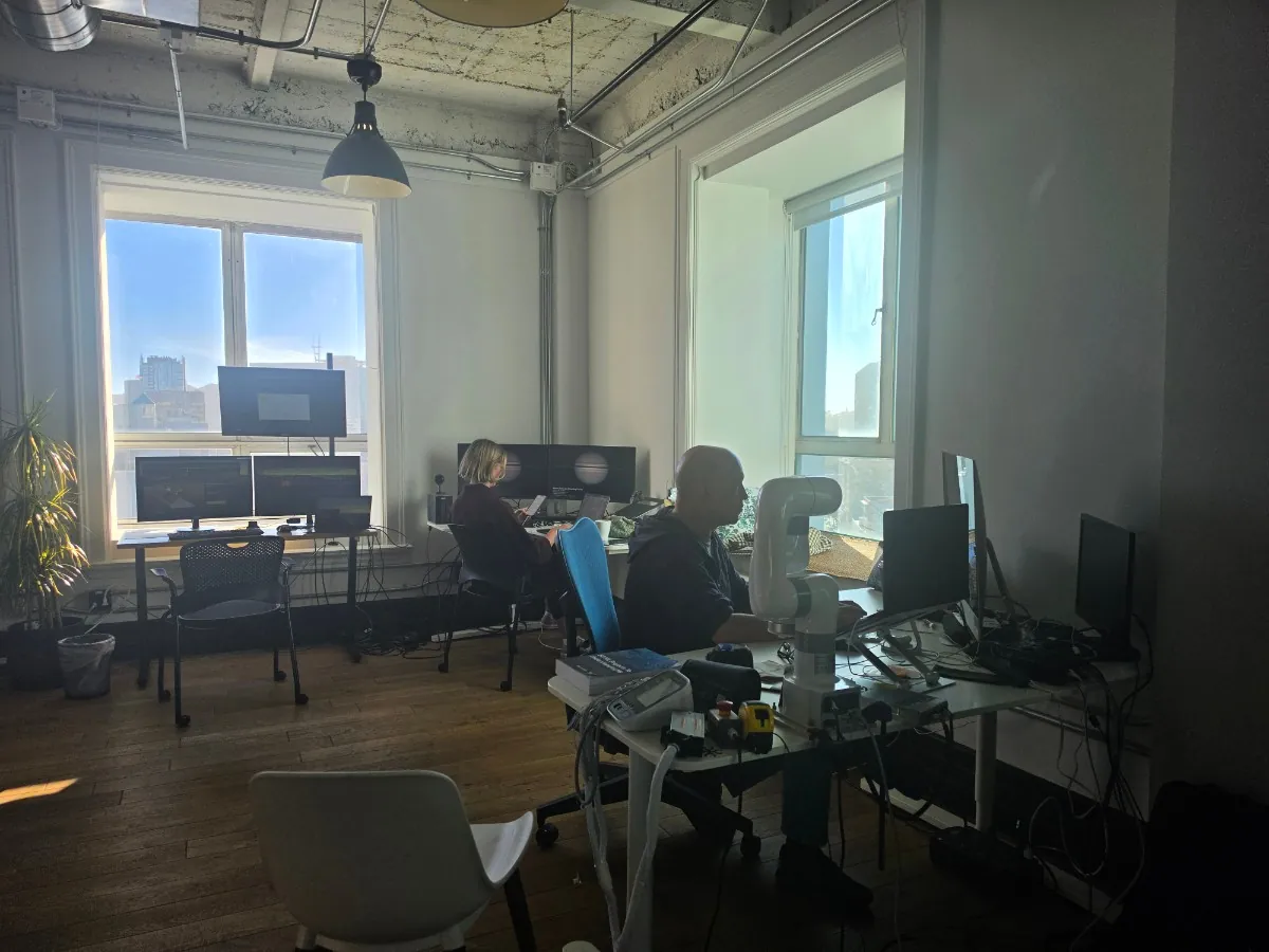 Researchers at their workstations inside the Other Minds Laboratory, with a robotic arm on the bench and the city visible through tall windows.